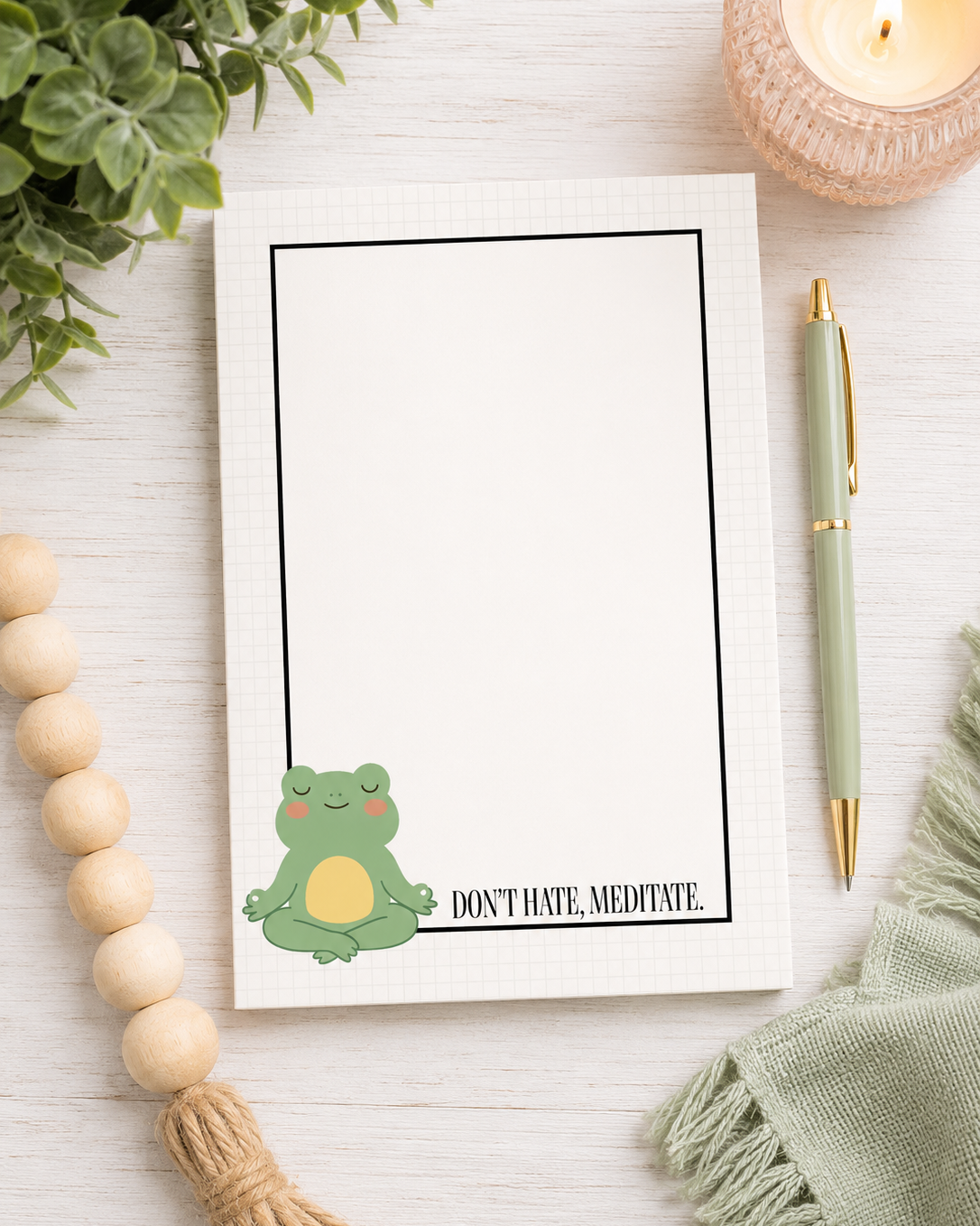 A white notepad with a black border and a cute green meditating frog illustration sitting at the bottom left, next to the text 'DON'T HATE, MEDITATE.' The notepad is styled in a flat-lay on a white wood background with a green pen, a lit candle, wooden beads, and a green cloth.