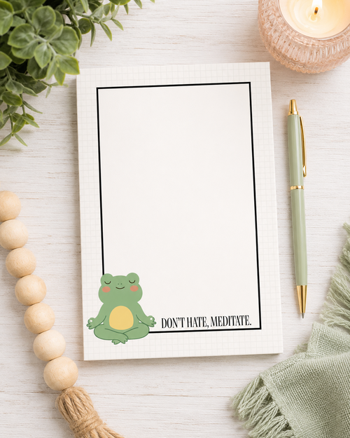 A white notepad with a black border and a cute green meditating frog illustration sitting at the bottom left, next to the text 'DON'T HATE, MEDITATE.' The notepad is styled in a flat-lay on a white wood background with a green pen, a lit candle, wooden beads, and a green cloth.