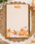 A 4x6 fall themed notepad with Notes at the top and watercolor pumpkins and flowers at the top placed on a wicker placemat with pen.
