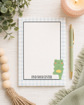 A flat-lay of a notepad with a light blue and white gingham border, featuring a cute green frog meditating illustration and the text 'FIND YOUR CENTER'. The notepad is styled on a light wood background with a green pen, a candle, wooden beads, and a green scarf.