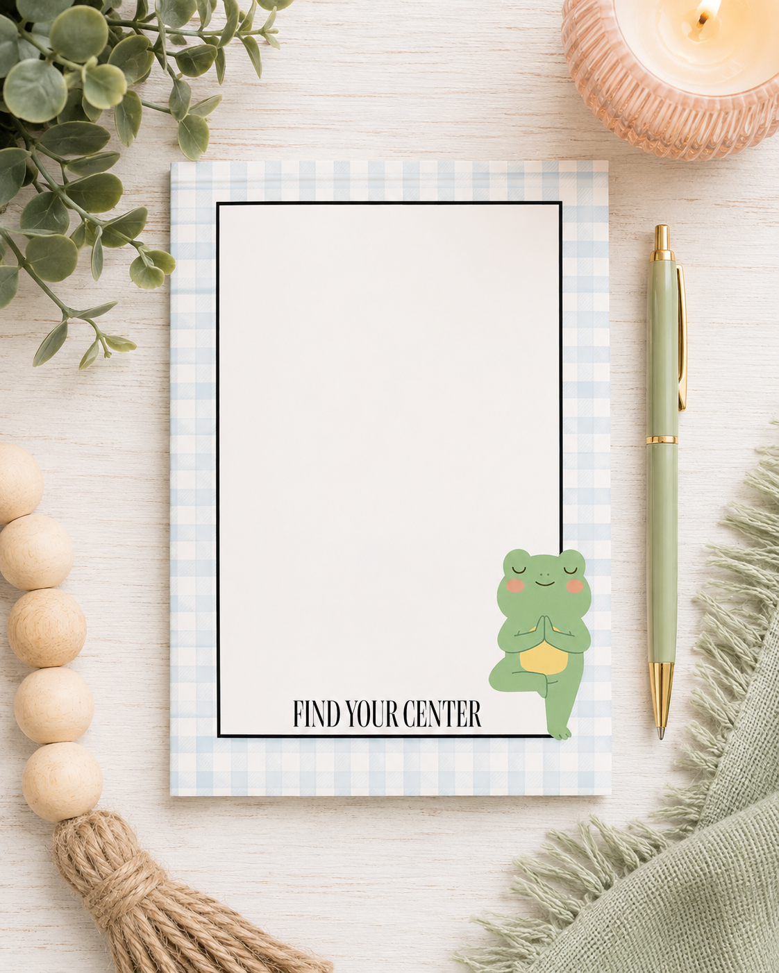 A flat-lay of a notepad with a light blue and white gingham border, featuring a cute green frog meditating illustration and the text 'FIND YOUR CENTER'. The notepad is styled on a light wood background with a green pen, a candle, wooden beads, and a green scarf.