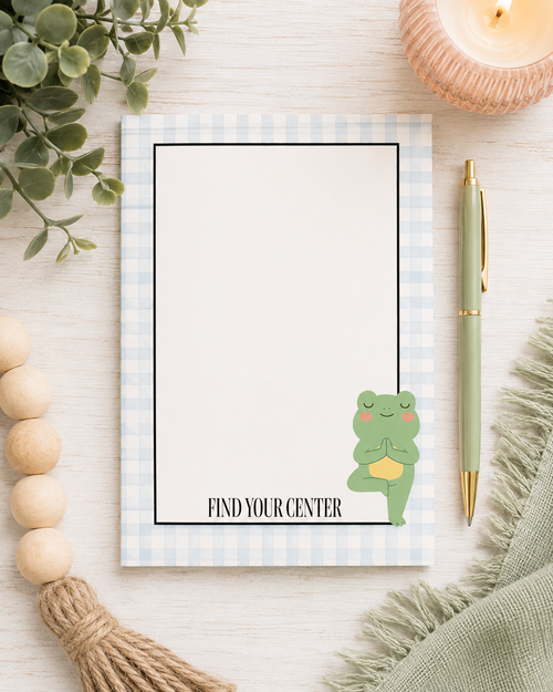 A flat-lay of a notepad with a light blue and white gingham border, featuring a cute green frog meditating illustration and the text 'FIND YOUR CENTER'. The notepad is styled on a light wood background with a green pen, a candle, wooden beads, and a green scarf.