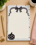 A 4x6 notepad with a black and white harlequin patterned border, featuring a black bow illustration at the top and a black pumpkin with pink hearts at the bottom, displayed on a light wood desk with a gold pen, binder clip, plant, and cup.