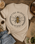 Kind Words Are Like Honey graphic Sand colored tee featuring a vintage-inspired bumblebee design and Proverbs 16:24, styled in a cozy cottagecore flat lay.