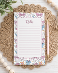 A decorative nurse themed notepad with lined writing space and the word 'Notes' at the top, styled on a braided jute placemat with a pink pen beside it on a white wood surface.