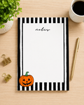 A Halloween-themed notepad with a black and white striped border and a cartoon jack-o'-lantern in the bottom-left corner, styled on a light wood desk with a gold pen, binder clips, a plant, and a cup of coffee.