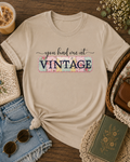 Sand Shirt with “You Had Me at Vintage” graphic tee styled in a cozy flat lay with iced coffee, denim shorts, sunglasses, crochet accessories, and vintage-inspired thrifted finds.