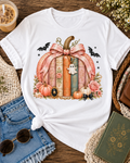 A white short-sleeve T-shirt laid flat on a rustic wooden surface, styled with jeans, sunglasses, an iced coffee, flowers, and a book. The shirt features a large autumn-themed graphic of stacked books forming a pumpkin shape, tied with a pink bow, with pumpkins, roses, bats, and a small ghost charm.