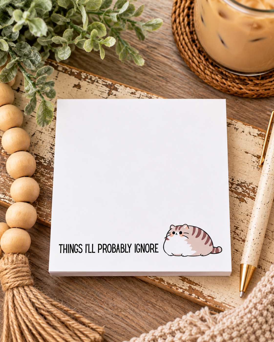 A white notepad with the printed phrase 'THINGS I'LL PROBABLY IGNORE' and a small cartoon cat illustration, styled on a rustic wooden surface with beads, greenery, a drink, and a pen.