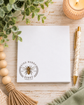 A white notepad with a bee logo and the words 'KIND WORDS  ARE LIKE HONEY' lies on a light wood surface, styled with a pen, candle, beads, leaves, and fabric.
