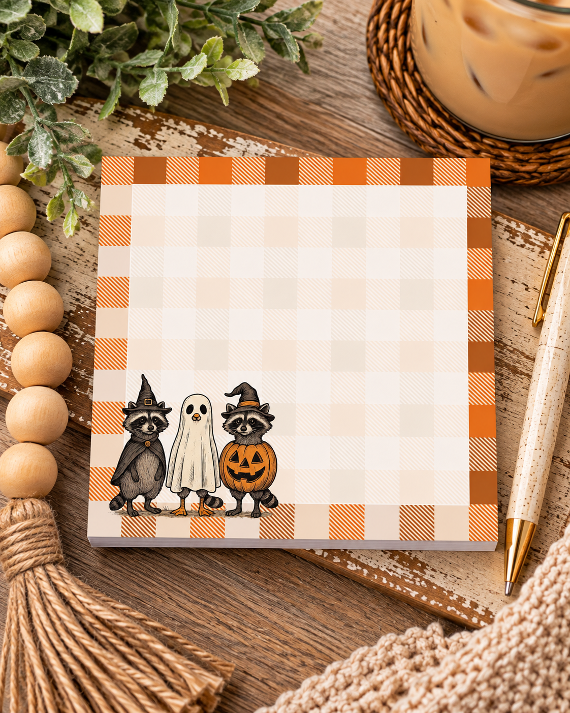 Square Halloween notepad with orange and brown plaid featuring two costumed raccoons and a ghost-costumed duck at the bottom layered on a wood table with wood beads, pen and iced coffee drink. 