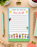 A personalized desk notepad with a light blue border, featuring the text 'FROM THE DESK OF Mrs. Smith' in red script, surrounded by colorful cartoon children and kites.