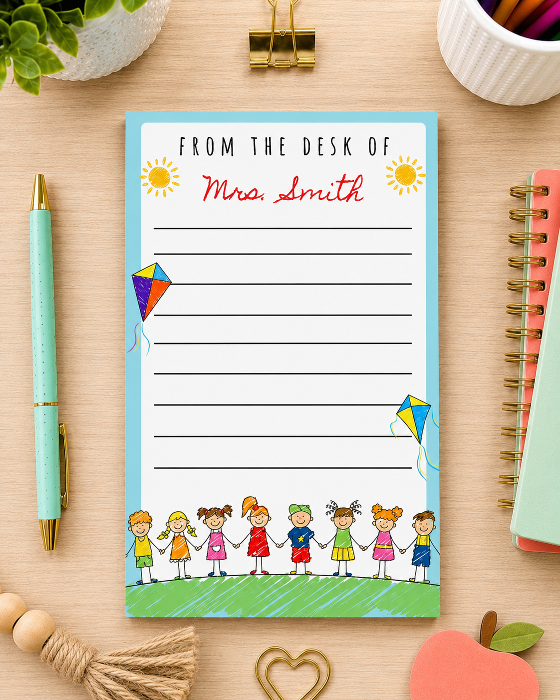 A personalized desk notepad with a light blue border, featuring the text 'FROM THE DESK OF Mrs. Smith' in red script, surrounded by colorful cartoon children and kites.